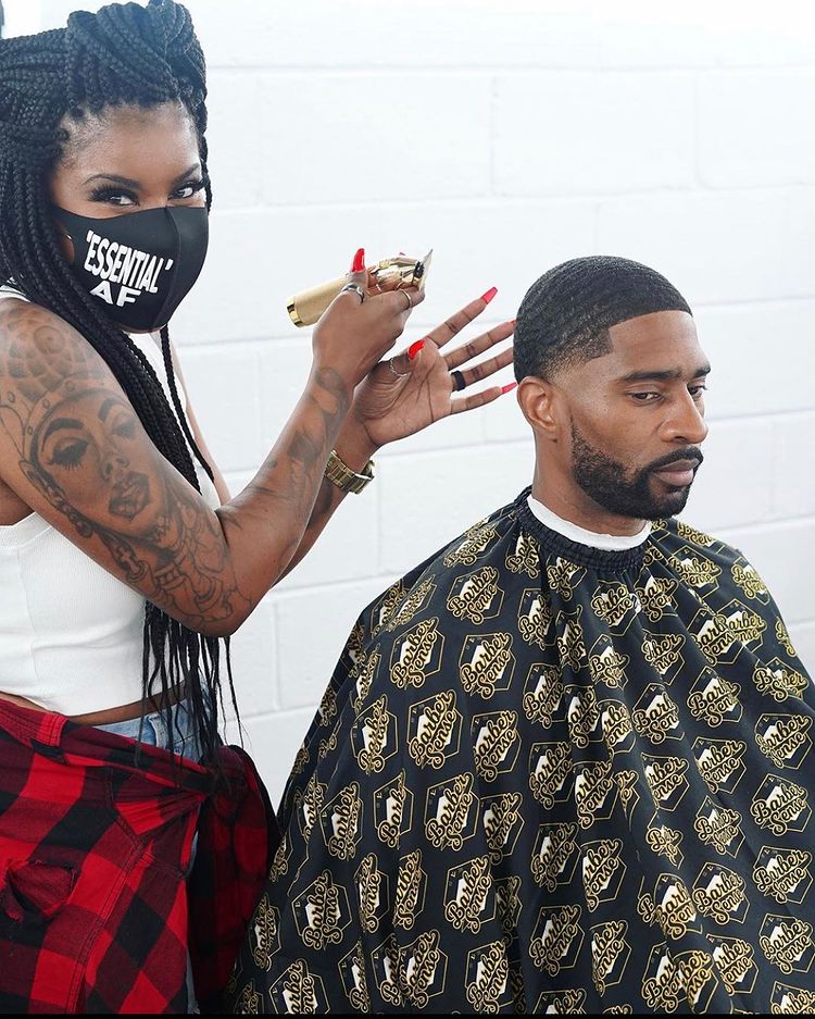 A Black woman barber wearing a mask that reads "essential AF" while at work.